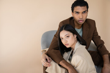 african american man in brown blazer embracing brunette asian woman and looking at camera isolated...