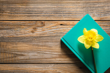 Green book and yellow narcissus flower on wooden background, top view.