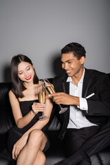 happy interracial couple in black elegant attire clinking champagne glasses on leather couch on grey background.