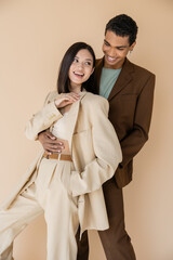 stylish african american man embracing carefree asian woman in ivory suit isolated on beige.