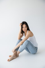 full length of barefoot asian woman in white tank top and jeans sitting and looking at camera on grey background.