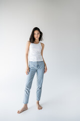 full length of barefoot and slim asian woman in tank top and jeans standing on grey background.