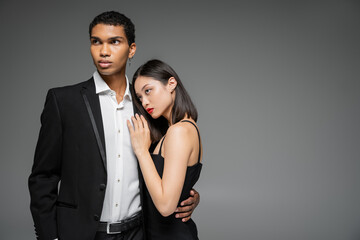 sensual asian woman in strap dress leaning of elegant african american man in black blazer isolated on grey.