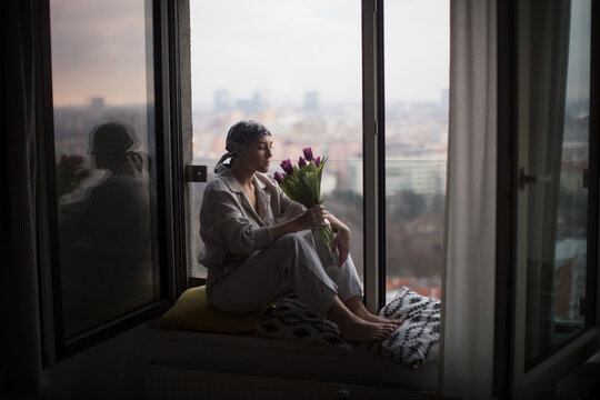 Young Woman With Cancer Sitting In A Window And Smelling Flowers.