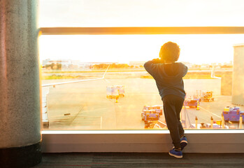 Young boy looking out of the window at an airport watching the planes at sunset. View from behind. Waiting for his flight on vacation