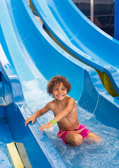 Smiling little diverse boy sliding down a waterslide at an outdoor waterpark on a summer day. Cute African American boy having fun on a warm sunny day.