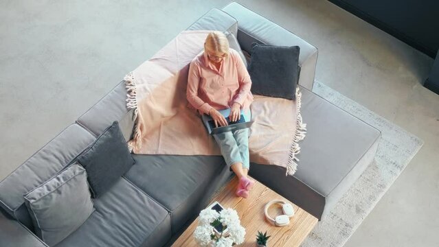 Top view of beautiful woman in casual clothes sitting on soft big gray sofa in living room and working on laptop typing on keyboard. Comfortable interior and remote work.