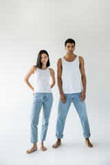 full length of young and barefoot multiethnic couple posing in white tank tops and blue jeans on grey background.