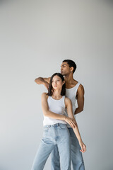 young african american guy touching neck of asian woman in white tank top and jeans isolated on grey.