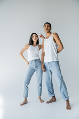 full length of barefoot interracial couple in blue jeans and white tank tops looking at camera on grey background.