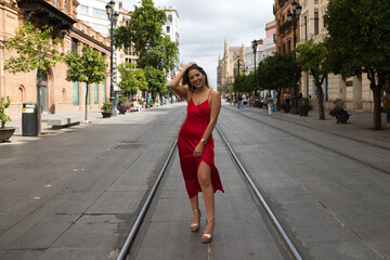 beautiful young woman in a red silk dress stands between the tram tracks on an avenue in seville. The model poses for the photo shoot. Fashion and beauty concept.