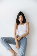 Fototapeta na wymiar brunette asian woman in blue jeans and white tank top sitting and looking at camera on grey background.