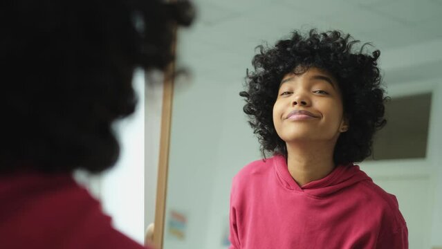 Love yourself. Beautiful young smiling african american woman dancing touching curly hair enjoying her mirror reflection. Black lady looking at mirror looking confident and happy. Self love concept