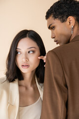 brunette asian woman looking away near african american man in brown blazer isolated on beige.