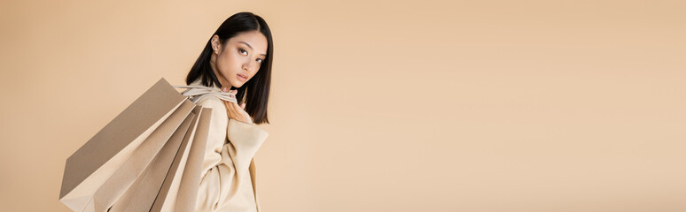 brunette asian woman in trendy blazer looking at camera while holding shopping bags isolated on...