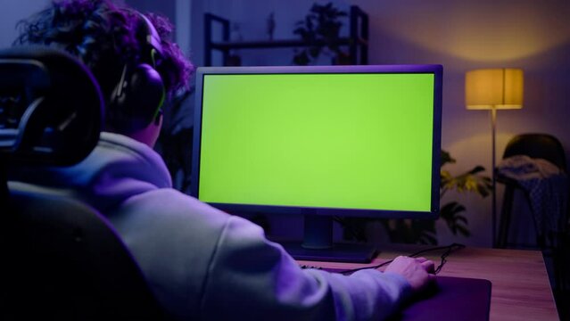 Young gamer in headphones plays video games late at night, sitting at laptop with chroma key screen