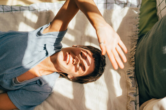 A woman covers her face with her hands from the bright sun rays while lying on a bed near the window
