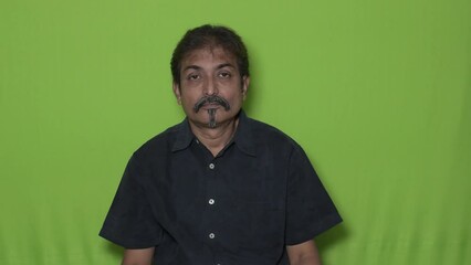 The well-dressed Indian man wearing a black shirt is indicating something to the top direction with his right hand's index fingers while standing against a green screen background