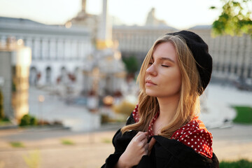 Fashion portrait of adorable stylish blonde young caucasian girl in beret and polka dot dress posing outside with urban view at sunset. Elegant concept. High quality image