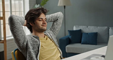 Young positive relaxed caucasian guy leaning back in his chair after finishing working on his laptop computer at home. Successful freelancer or remote worker on lockdown 