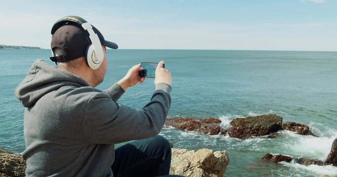 Man in White Headphones Listens to Music Photographs Nature on a Smartphone While Sitting on the Shore of a Rocky Ocean. 