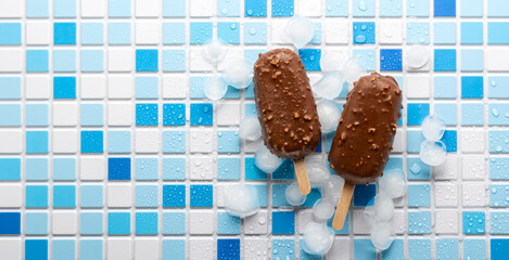 Two ice cream popsicles in chocolate nut glaze on ice, on a white and blue tile. Summer dessert, eskimo top view. Banner with copy space. - obrazy, fototapety, plakaty