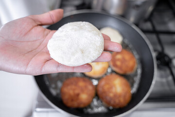A hand is holding a potato cake before frying it in a sauce pan with vegetable oil