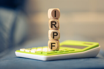 A sigla IRPF escrita em dados de madeira que estão empilhados sobre uma calculadora. Imposto de...