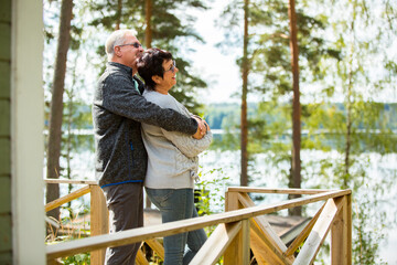 Mature couple standing on porch and hugging. They are smiling and looking at the beautiful view - forest and lake. Happy senior couple embracing each other on the wooden terrace of the house. 