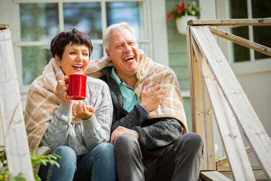 Mature couple sitting on porch and drinking hot tea from mug. They are smiling and laughing, wrapped in a blanket. Happy senior couple embracing each other on the wooden terrace of the house. 