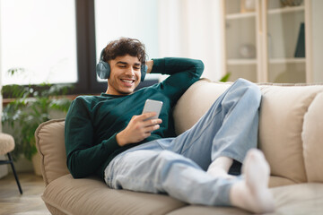 Guy Using Phone Wearing Headphones Lying On Sofa At Home