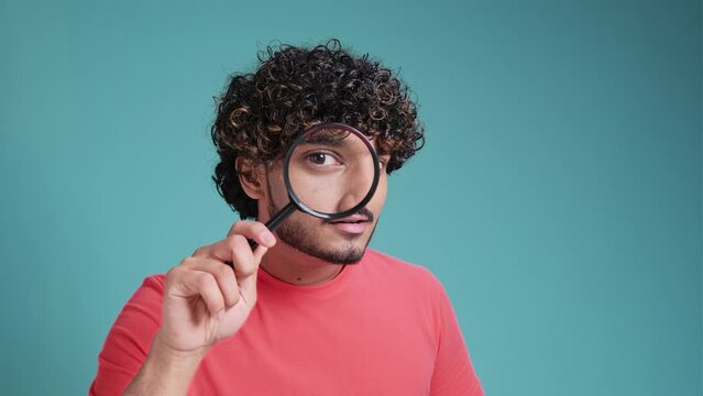 Serious young latin hispanic male detective holding magnifying glass by right eye while standing on blue studio background