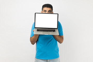 Portrait of shy young adult man wearing blue T- shirt standing hiding half of his face behind laptop with empty display, looking at camera. Indoor studio shot isolated on gray background.