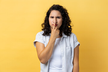 Portrait of woman with dark wavy hair looking with incredulous suspicious gaze and touching nose,...