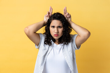 Portrait of strict serious woman with dark wavy hair showing bull horn gesture with fingers over...