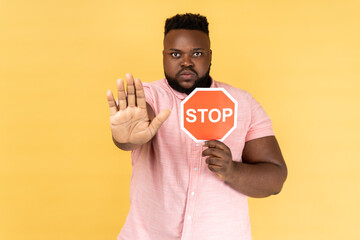 Portrait of man wearing pink shirt holding red stop sign, looking at camera with ban palm gesture,...