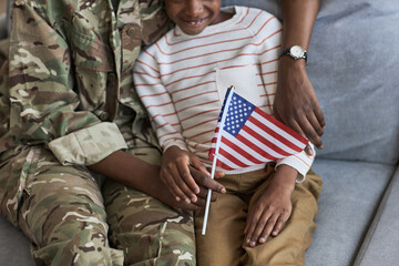 Close-up of little child sitting with american flag on sofa and supporting her mom in military uniform