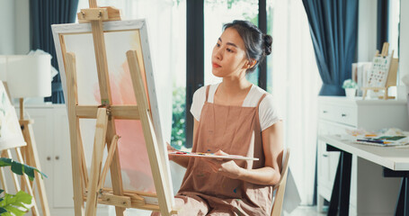 Happy youth Asia Female wear apron holding palette color use brush painting on canvas create artwork in cozy workshop at home. Contemporary Painter Abstract Modern Art, Creativity and people concept.