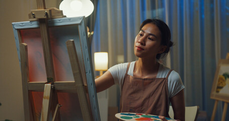 Happy youth Asia Female wear apron holding palette use brush painting on canvas create artwork cozy workshop in home at night. Contemporary Painter Abstract Modern Art, Creativity and people concept.