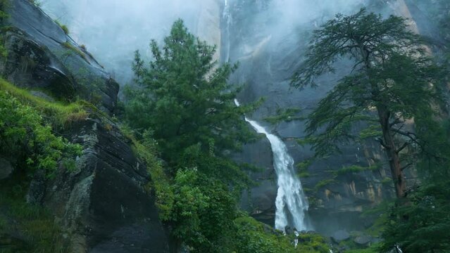 Slow motion shot of flowing Jogini waterfall covered with lush green trees in mountains of Manali, Himachal Pradesh, India. Tranquil View of Jogini falls with rain and foggy weather during monsoon. 