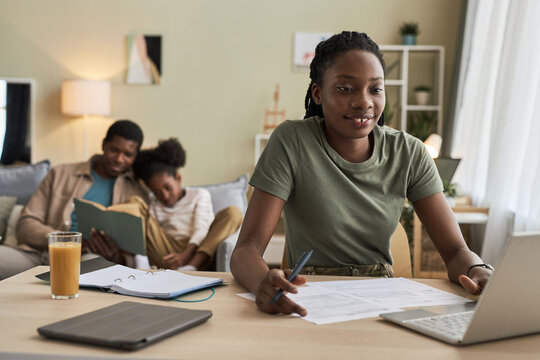 African American woman working online on laptop at home while father reading a book with child in the background - Powered by Adobe