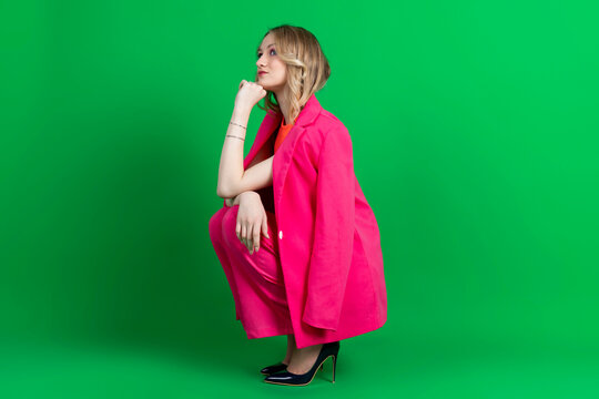 Young curly blonde dressed in pink blazer and pants, crouches and thoughtfully looking up. Isolated on green background.