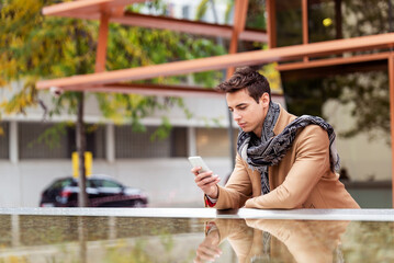 Portrait of stylish handsome young man with coat standing outdoors and leaning on wall using a smartphone
