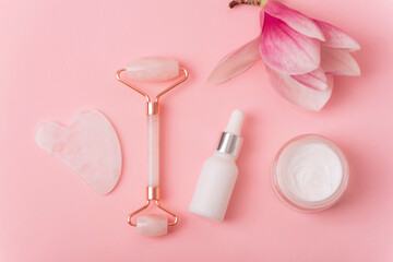 Rose quartz crystal facial roller and gua sha scraper, face serum or organic cosmetic oil, magnolia flower on pink background. Facial massage kit for lifting therapy. Top view, flat lay