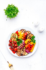 Grilled vegetables: paprika, zucchini, eggplant, mushrooms, tomatoes and onion on plate, white table background, top view