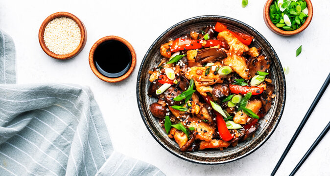 Stir fry chicken fillet, red paprika, mushrooms, green chives and sesame seeds in ceramic bowl Asian cuisine dish. White kitchen table background, top view