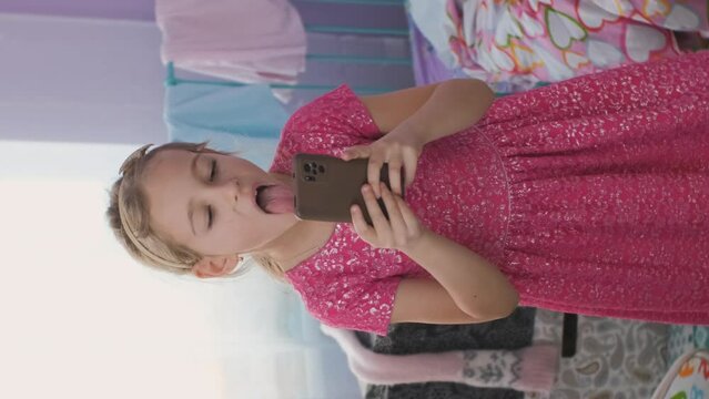 Vertical video. Little girl in pink dress grimacing while taking selfies on her smart phone in messy room, slow motion