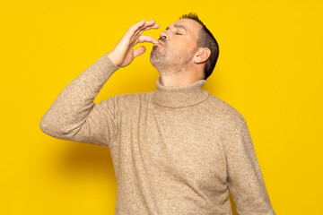 Bearded Hispanic man wearing a beige turtleneck smacking his lips and licking his fingers after devouring an exquisite delicacy, isolated on yellow studio background. - obrazy, fototapety, plakaty