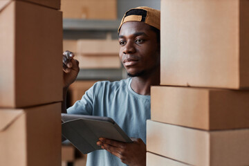 Selective focus on young man with tablet standing between stacks of boxes and pointing at one of...