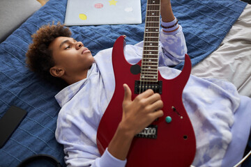 Relaxed African American teenage boy in hoodie lying on comfortable bed and playing electric guitar...